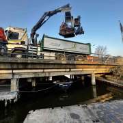 Rupsvoertuig + Maaiarm wat opgetild voor werkzaamheden aan een talud vanaf het water, op een ponton.