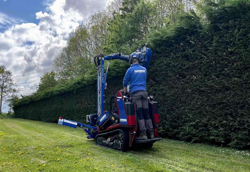 Smal rupsvoertuig, uitgerust met een maaiarm voert snoeiwerkzaamheden uit aan een conifeer.